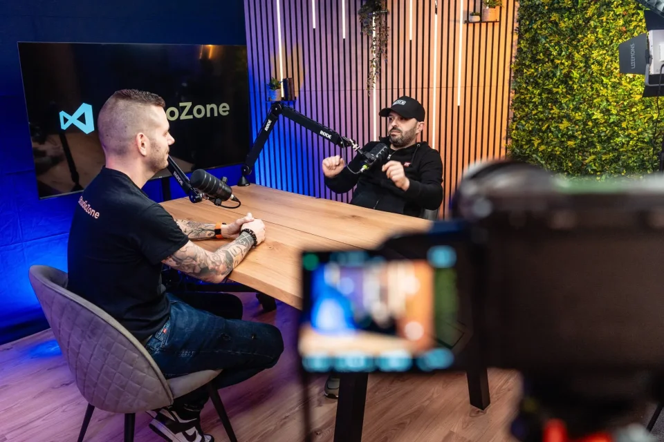 Podcast felvétel a Zóne stúdióban: műsorvezető az asztalnál, 55 colos StudioZone kijelző, kamera előtérben