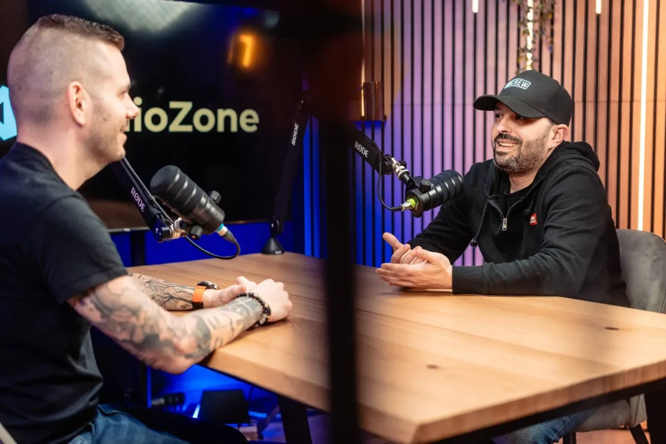 Két beszélgető a Zóne stúdió asztalánál Rode mikrofonokkal, háttérben StudioZone képernyő és mohafal
