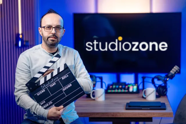 Szemüveges műsorvezető StudioZone csapótáblával, háttérben studiozone kijelző és keverő